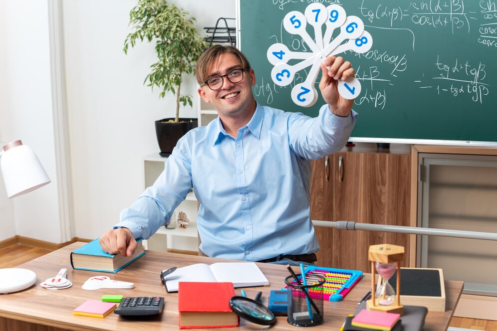 profesor sexo masculino joven gafas mostrando placas matricula explicando leccion sonriendo sentado escritorio escuela libros notas frente pizarra aula 141793 102238